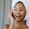 Young woman with towel on head, smiling and applying skincare product, promoting morning beauty routine.