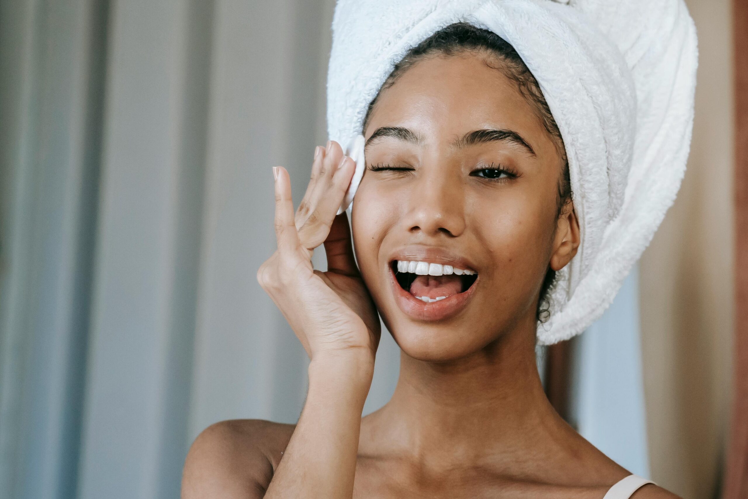 Young woman with towel on head, smiling and applying skincare product, promoting morning beauty routine.
