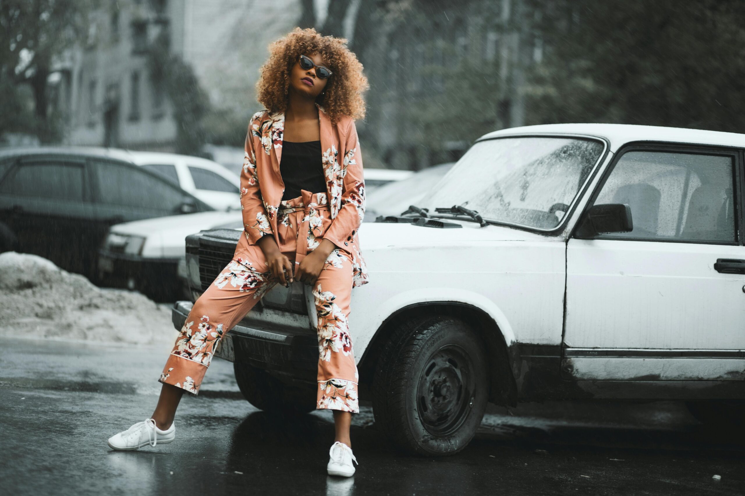 Stylish woman in floral suit posing against a classic car on a rainy day.